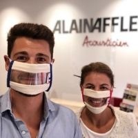 Selfie d'un jeune homme et d'une jeune femme portant des Masques Sourire devant un mur affichant le logo Alain Afflelou Acousticien 